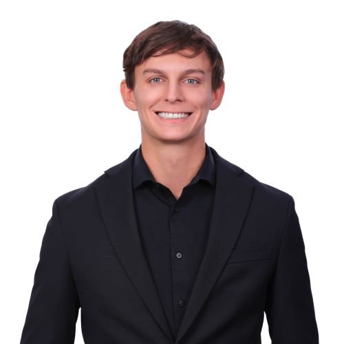 Portrait of a smiling young man with short brown hair wearing a black suit and black dress shirt against a white background.