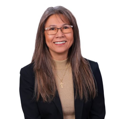 Portrait of a woman with long brown hair, glasses, wearing a black blazer over a beige top, smiling against a white background.